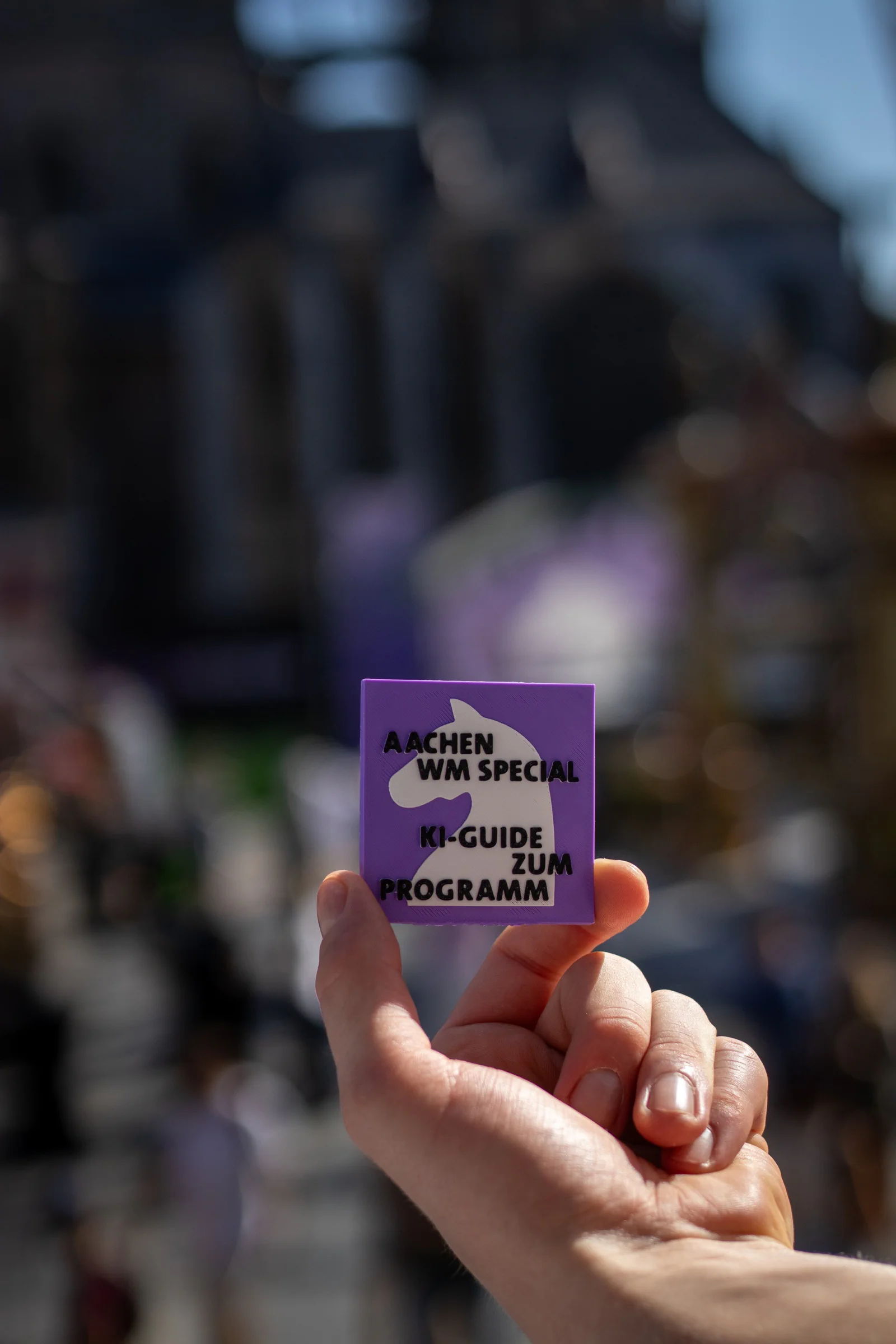 Eine Hand hält einen violetten 3D-gedruckten KI-Guide zum Programm vor der unscharfen Aachener Innenstadt